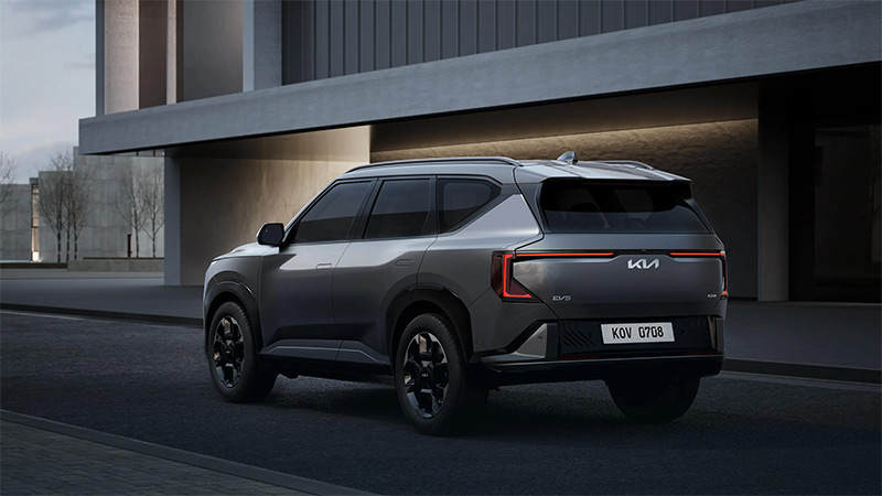 Šedý model vozu Kia EV5 parkující na cestě před moderní budovou - boční pohled ze zadu