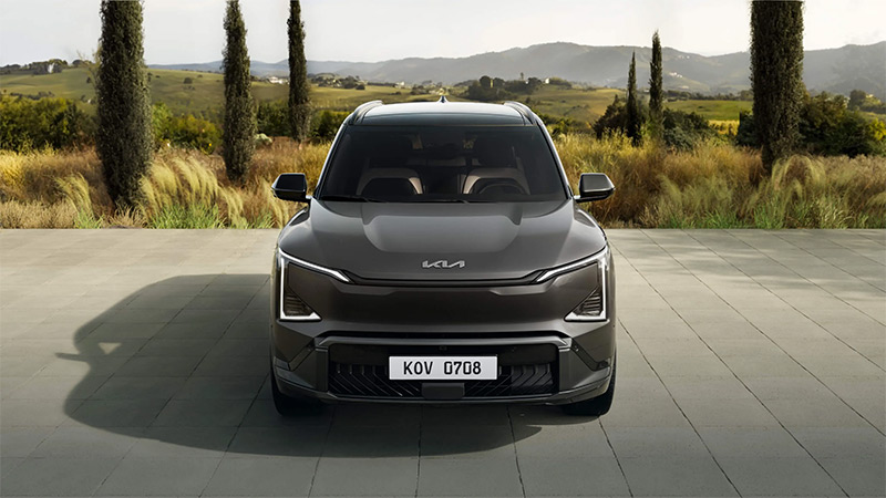 Šedy model vozu Kia EV5 - přední pohled na zaparkovaný vůz se zapnutými světly, v pozadí příroda