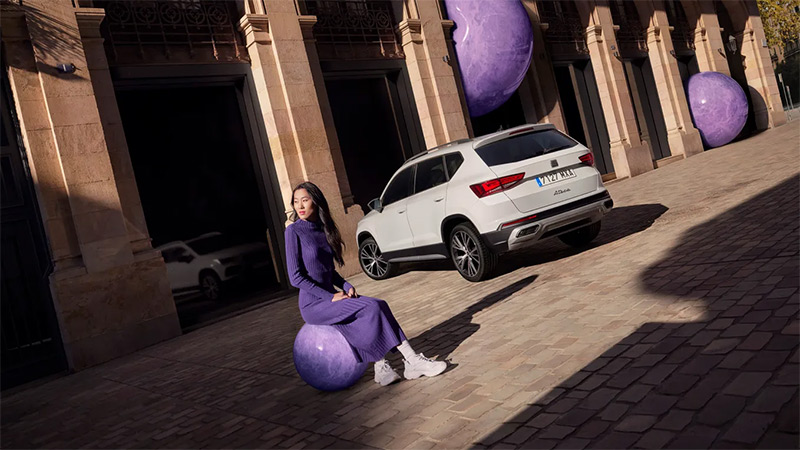 6ena sedí na fialovém míči vedle zaparkovaného bílého vozu SEAT Ateca