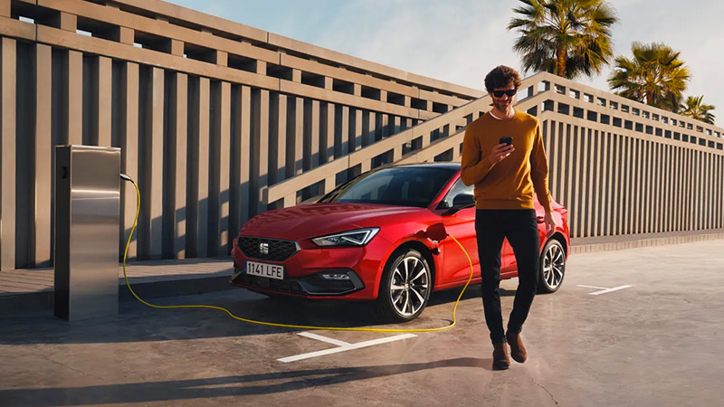Červený SEAT Leon stojí u nabíjecí stanice a dobíjí se, muž odchází s telefonem v ruce